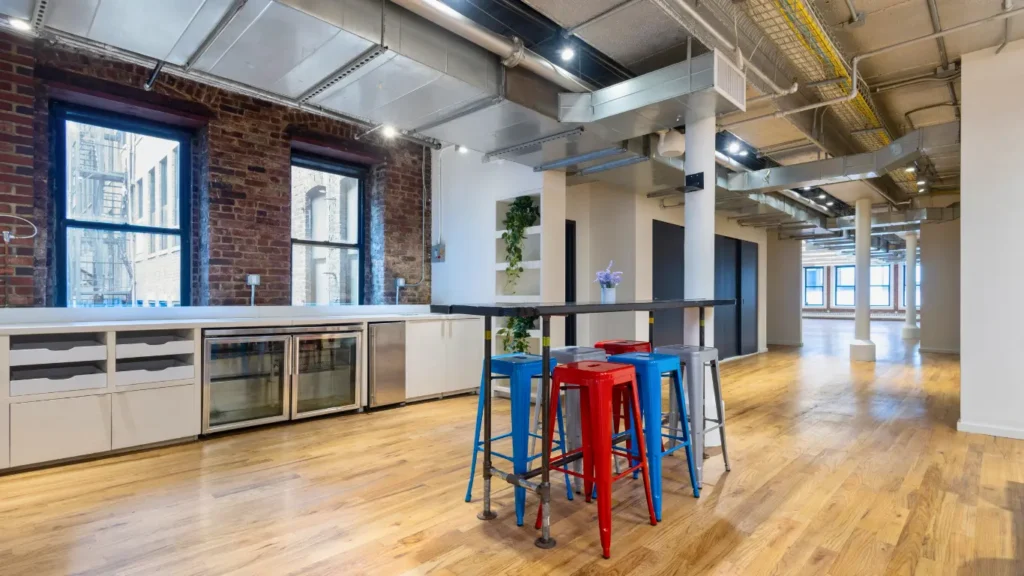 Spacious coworking lounge and break room with high ceilings, exposed ductwork, and colorful bar stools around a tall communal table.