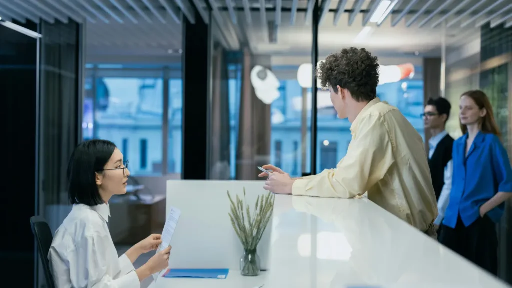 Receptionist speaking with a visitor at a modern coworking space front desk while other guests wait to book meeting rooms.