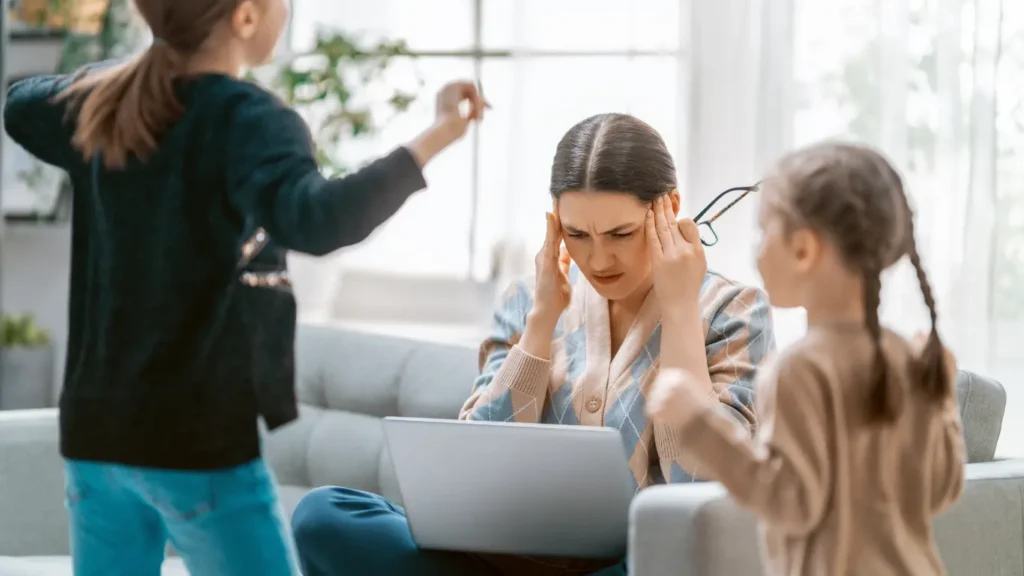 Stressed remote worker on a sofa with a laptop, rubbing their temples while two energetic children play noisily nearby.