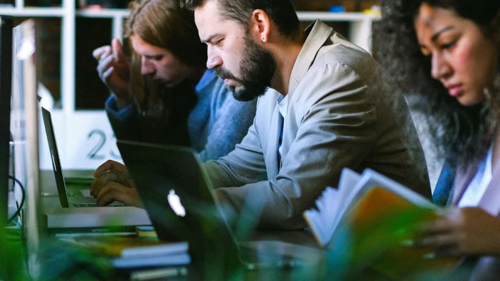 Coworkers using laptops at a shared workspace table.