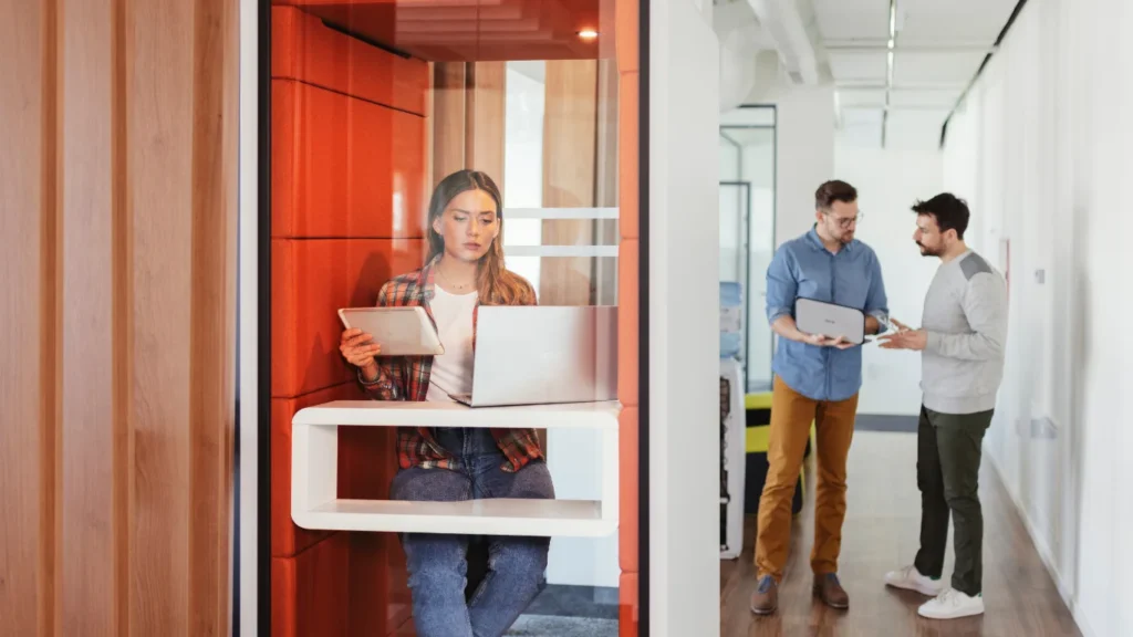 Woman working in a private office booth while two colleagues talk nearby.
