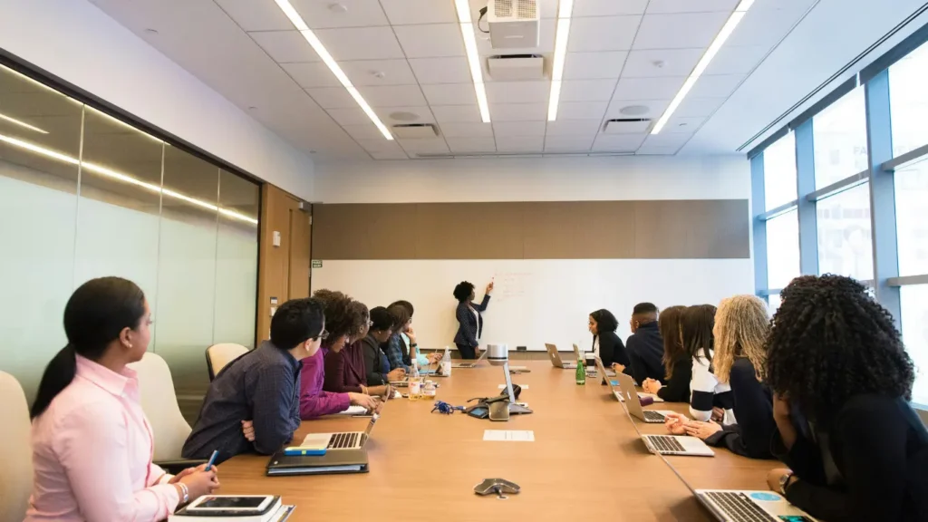 Large conference room meeting with people seated around a long table facing a presenter.